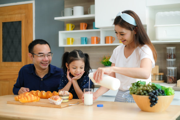 可爱的小女孩和亚洲爸爸妈妈把牛奶从瓶子倒进杯子里给女儿在厨房当早餐喝。图片下载