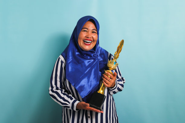 一位戴着头巾的亚洲中年妇女兴奋地拿着一个金色奖杯，孤立在蓝色的背景上图片下载