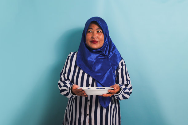 一位戴着头巾的中年亚洲妇女沉思着要吃什么菜，孤立在蓝色的背景上图片下载