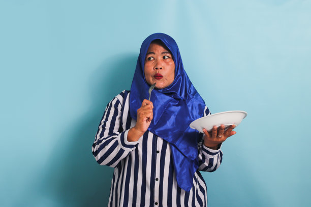 戴着头巾的中年亚洲妇女拿着白色的盘子和勺子，在蓝色的背景上显得很孤立图片下载