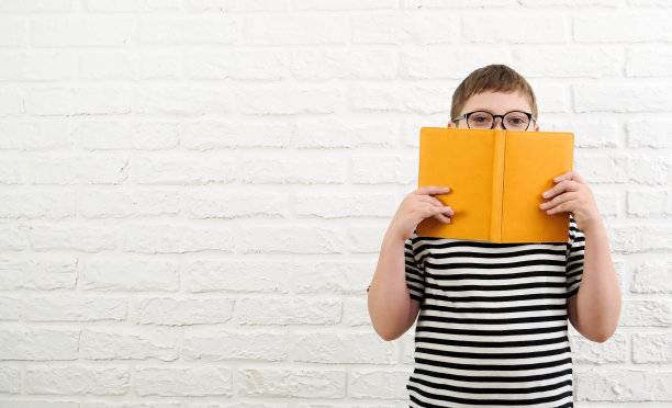 一个可爱开朗的男孩，穿着条纹t恤，戴着眼镜，拿着一本黄色的书，半脸贴在白砖背景上。图片下载