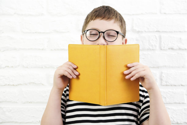 一个可爱开朗的男孩，穿着条纹t恤，戴着眼镜，拿着一本黄色的书，半脸贴在白砖背景上。图片下载