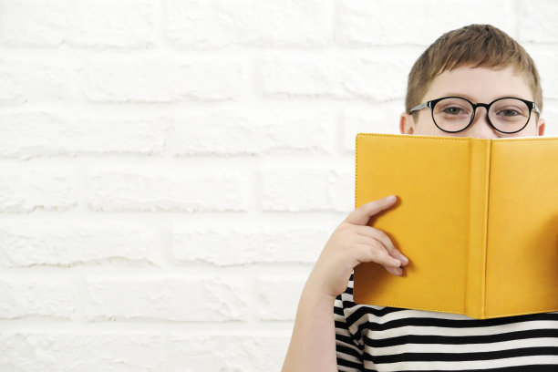 一个可爱开朗的男孩，穿着条纹t恤，戴着眼镜，拿着一本黄色的书，半脸贴在白砖背景上。图片下载