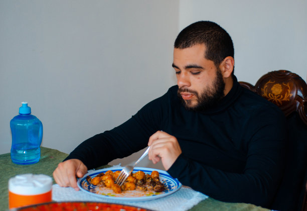 一个穿着黑色休闲上衣的男人坐在餐桌旁吃着一顿有肉和土豆的午餐，手里拿着叉子看着食物。即食食品图片下载