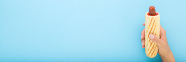 年轻的成年女子手拿着新鲜的法国热狗，在浅蓝色的桌子背景上涂着红色的番茄酱。柔和的颜色。宽横幅。特写镜头。快餐。空白位置用于文本。自顶向下视图。图片下载