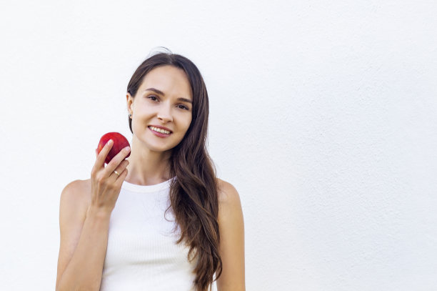 一个年轻迷人的女人，棕色长发，白色上衣，白色背景，拿着油桃或桃子，微笑着，孤立的拷贝空间。美丽健康的生活理念图片下载