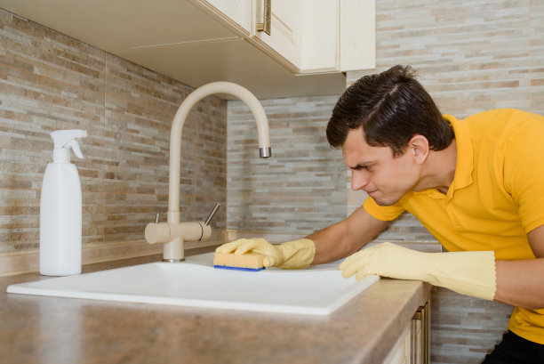 穿着黄色t恤、戴着黄色手套的年轻人正在用清洁喷雾和海绵清洁厨房的洗脸盆水槽。图片下载