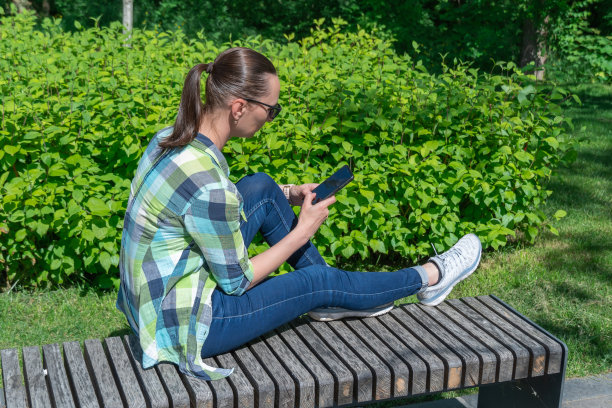 一个女人坐在公园的长椅上，翻看着手机的显示屏。图片下载