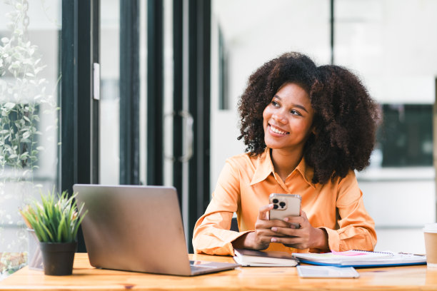 黑人女商人一边用笔记本电脑工作，一边用智能手机。微笑的非洲裔美国妇女在办公室用手机应用玩游戏、网上购物和叫外卖。图片下载