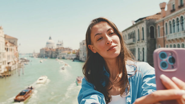 可爱的快乐女孩，棕色长发，穿着蓝色衬衫，站在威尼斯运河的背景上用手机自拍图片下载