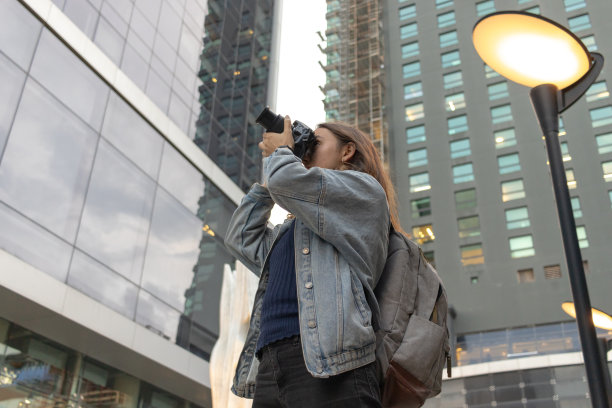 年轻的女摄影师穿着休闲服，背着摄影师的背包，在被建筑物包围的街道上拍照。图片下载