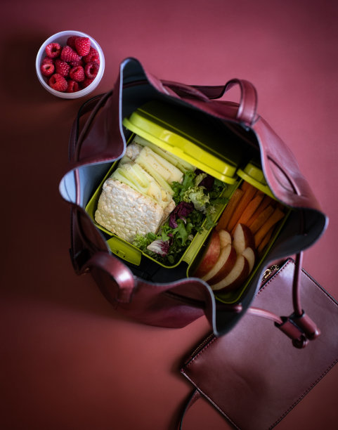 午餐盒里有三明治、胡萝卜条、苹果、生菜和覆盆子，以酒红色为背景，还有一个女人的手提包和钱包。健康食品的概念。俯视图图片下载