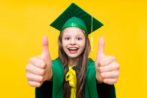 一个戴着毕业帽的女孩竖起大拇指。一个穿着大师服装的聪明孩子。黄色孤立背景。图片下载