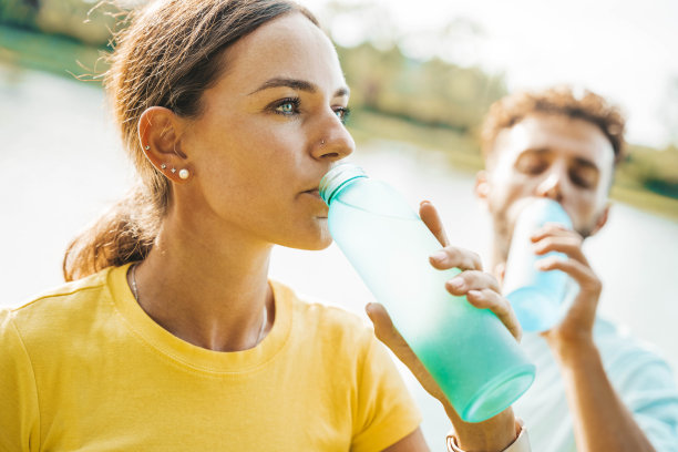 年轻女子和男子在跑步后喝着热水瓶。一对美丽的夫妇在湖边的公园里锻炼后休息。健身、运动和健康理念图片下载