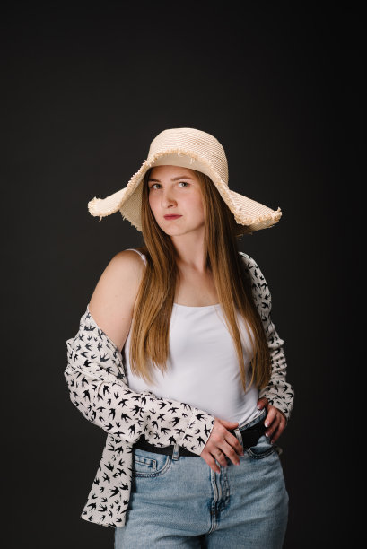 漂亮的女孩双手叉腰看着镜头。年轻的微笑的女人被隔离在墙上的复印空间。肖像中的女士穿着休闲时尚的服装，黑色背景上的帽子看起来很自信。照片。图片下载