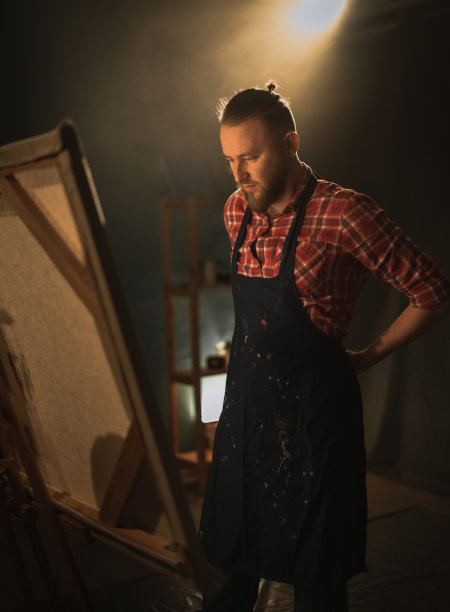 青年艺术家系着围裙，在黑暗的画室里准备用毛笔在画架上作画图片下载