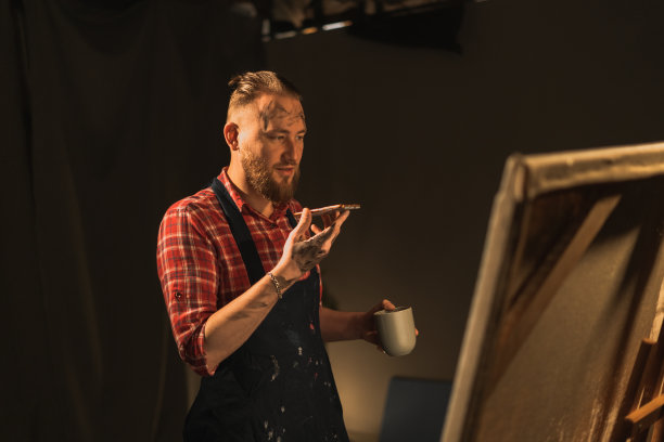 男艺术家站在他的现代创意工作室，用大画布，拿着免提和朋友打电话，用虚拟助手进行语音识别或请求图片下载