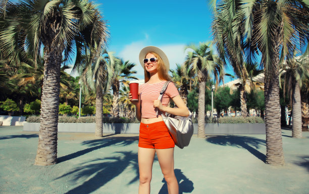 美丽的年轻女子在夏日公园拿着一杯咖啡，背着背包，戴着草帽，穿着短裤，背景是棕榈树图片下载