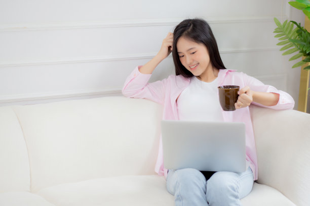 年轻的亚洲商业女性在家里用笔记本电脑工作，在客厅的沙发上喝咖啡，自由职业女孩坐在沙发上使用笔记本电脑，舒适放松，生活方式理念。图片下载