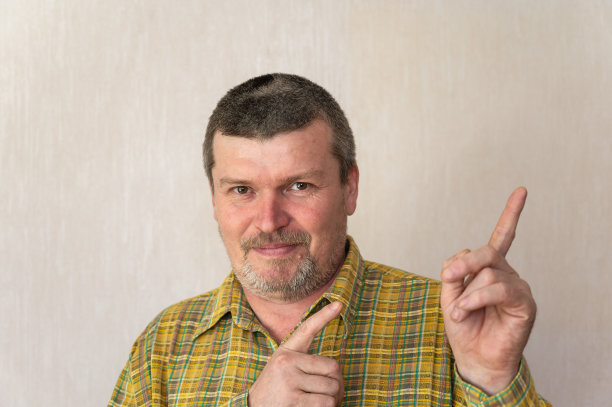 一个欢快的成年男子的肖像，在米色背景下穿着格子衬衫。一个头发花白的短发男子将食指向上指向一边。前视图。在室内。图片下载
