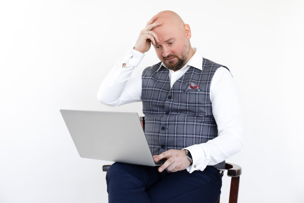 中年男子身穿灰色格子背心，蓝色牛仔裤，白色衬衫，坐在椅子上，拿着笔记本电脑。图片下载