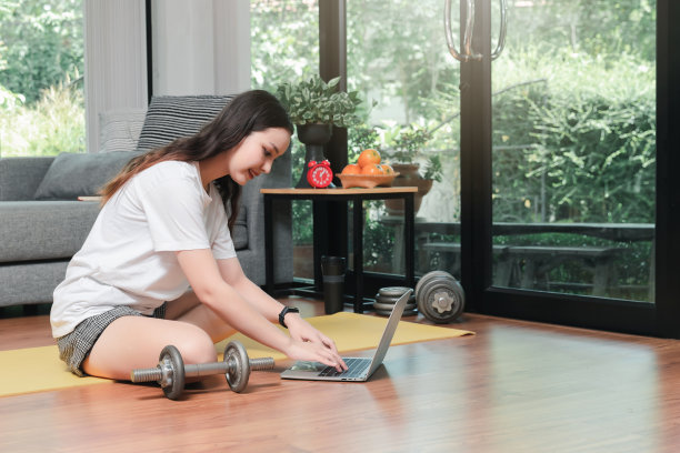 美丽的年轻女子与哑铃观看健身视频教程在笔记本电脑上，而坐在地板上的客厅。图片下载