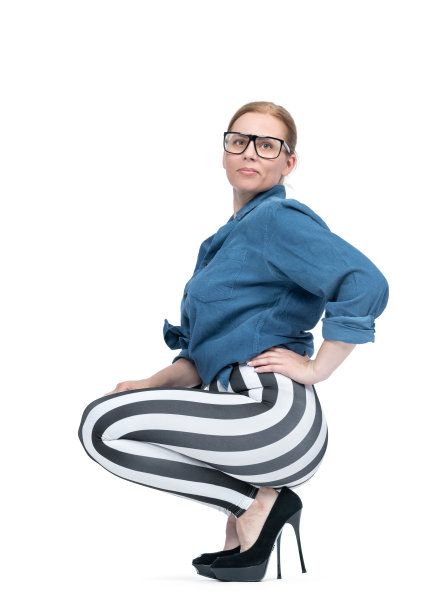 快乐的运动女性，戴着眼镜，穿着衬衫，条纹打底裤和细高跟鞋，蹲着。侧视图，孤立在白色背景上。图片下载
