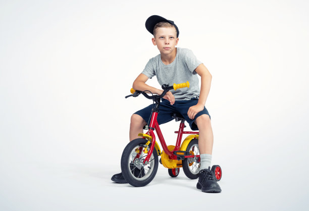 一个快乐的十几岁的男孩，穿着t恤、帽子和短裤，坐在一辆自行车上，背景是浅色的。文件包含到隔离的路径。图片下载