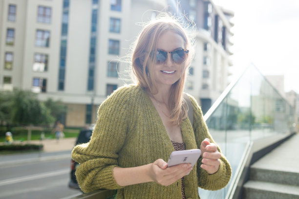 肖像微笑着年轻的高加索美女戴着太阳镜坐在城市外面看手机图片下载