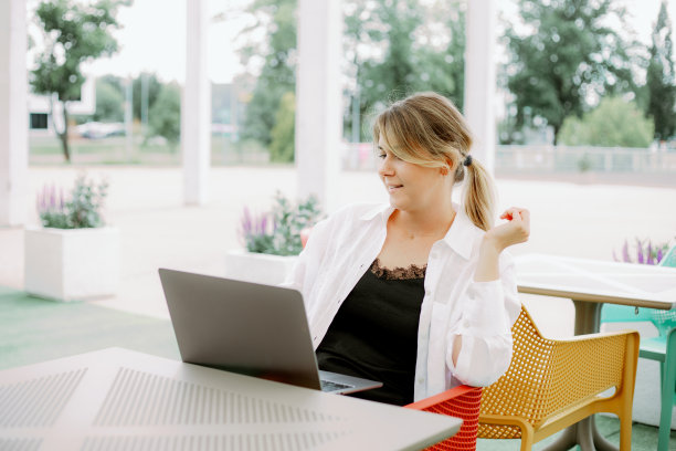 可爱的女人坐在桌子的椅子上，带着笔记本电脑，微笑着看着屏幕，在户外餐厅工作。图片下载