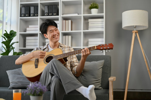一个开朗的亚洲人坐在客厅的沙发上唱着歌，弹着吉他。图片下载