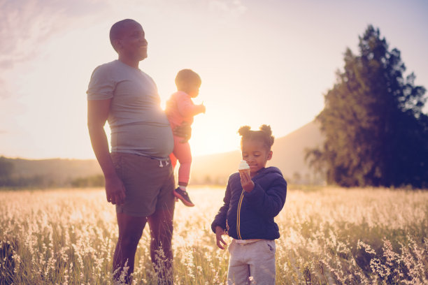 日落时分，一个小女孩与她的父亲和小妹妹在户外的草地上吃着冰淇淋图片下载