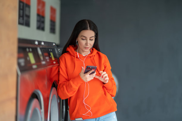 年轻女子用智能手机和耳机听音乐，在洗衣房等候。图片下载