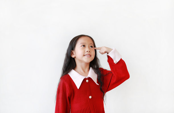 漂亮的小女孩穿着鲜红的裙子，用食指指着头，在白色的背景下抬起头思考。图片下载