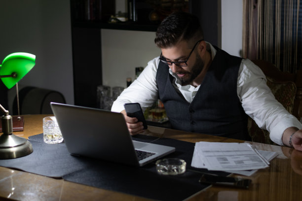 严肃英俊的商人，西装革履，倚在桌子上看图表。深夜工作的概念。工作到深夜为第二天做准备，完成他的工作。图片下载