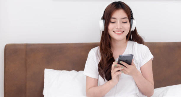 美丽的亚洲女性戴着耳机，从她们的智能手机上听音乐。在白色卧室的床上图片下载