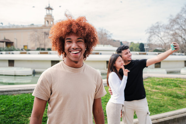 前景是一个快乐的非洲裔美国人，留着非洲发型，看着镜头，而他的朋友们正在自拍。年轻的学生男子对着背景微笑，一对夫妇正在拍照。少年男笑图片下载
