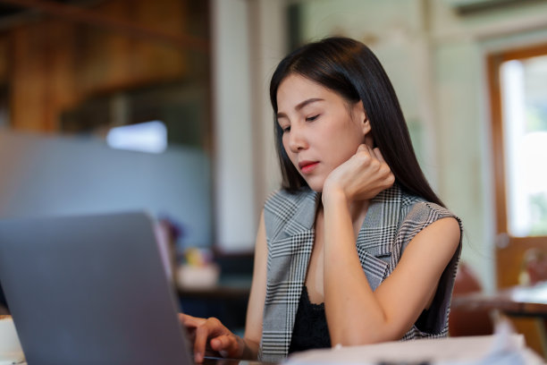 咖啡店的老板是一个漂亮的长发亚洲女人。无聊地坐着工作，把手放在下巴上在咖啡馆的桌子上使用笔记本电脑，检查桌子上的文件图片下载