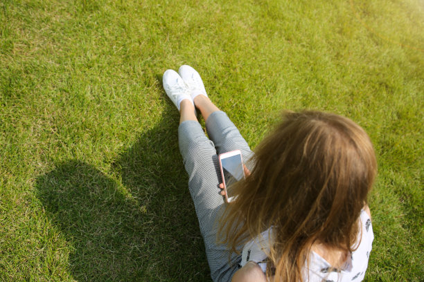 漂亮的时髦女孩坐在户外，时尚的商务女性在绿色的草坪上看智能手机。上网的学生。生活方式的放松。上班族。图片下载