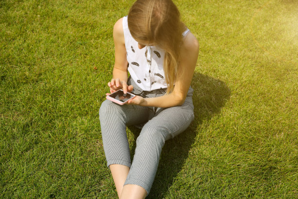 漂亮的时髦女孩坐在户外，时尚的商务女性在绿色的草坪上看智能手机。上网的学生。生活方式的放松。上班族。图片下载