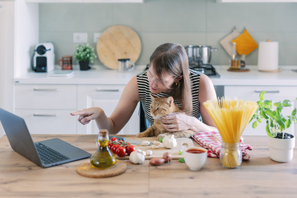 猫和它的主人穿着围裙在家里的厨房里一起做饭。家猫看着女孩如何给食物加盐。图片下载