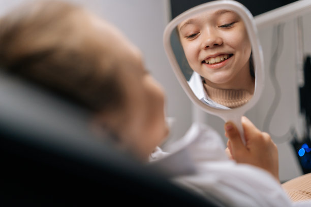 快乐可爱的小女孩病人对着镜子微笑，看着健康洁白的牙齿，享受牙科诊所的牙科保健设施。图片下载