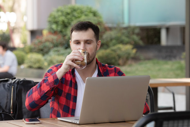 年轻的潮人在酒吧喝咖啡休息，他拿着杯子在笔记本电脑上打字。图片下载