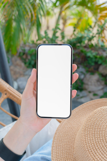 一个女人坐在游泳池旁边的椅子上，手里拿着空白屏幕的智能手机模型，暑假的概念。图片下载