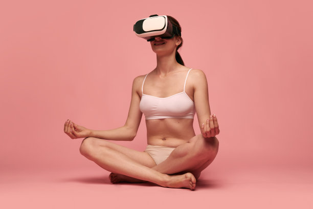 在线冥想。身材苗条的年轻女子穿着内衣，摆出莲花瑜伽的姿势，戴着vr眼镜，背景是粉红色的工作室图片下载