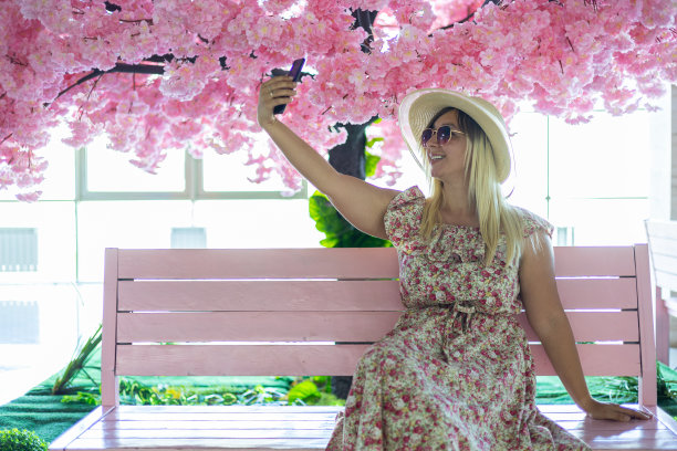 在购物中心，一名女子坐在人造樱花树下的长凳上，用智能手机自拍。在美丽的背景下，一名顾客在自拍留念。图片下载