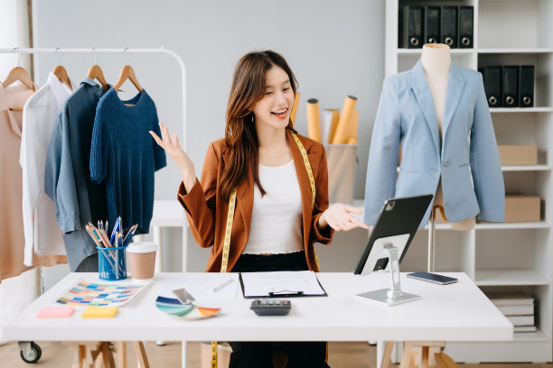 时装设计师女士在现代工作室里讲智能手机，用笔记本电脑和数码平板电脑，衣服挂在架子上图片下载