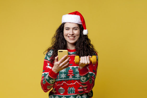 小女孩身穿圣诞服装，头戴圣诞帽，在黄色背景下练习健身，使用智能手机图片下载