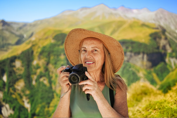 一位微笑快乐的老妇游客的肖像，她拿着相机，在群山的背景下摆姿势。在乡村草地上戴草帽的优雅老太太。积极退休人士概念图片下载