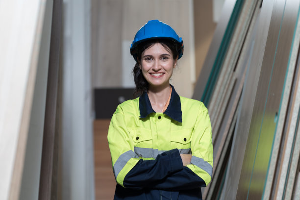 年轻女木匠的肖像，穿着安全制服和头盔在木工仓库储存车间工作。快乐微笑的木工女工在木材厂工作图片下载